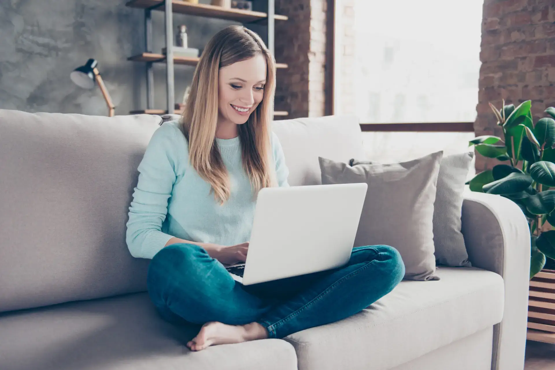 Portrait of positive, pretty, charming, domestic girl sitting on sofa having laptop on her legs, checking email, chatting with friend, using wifi internet