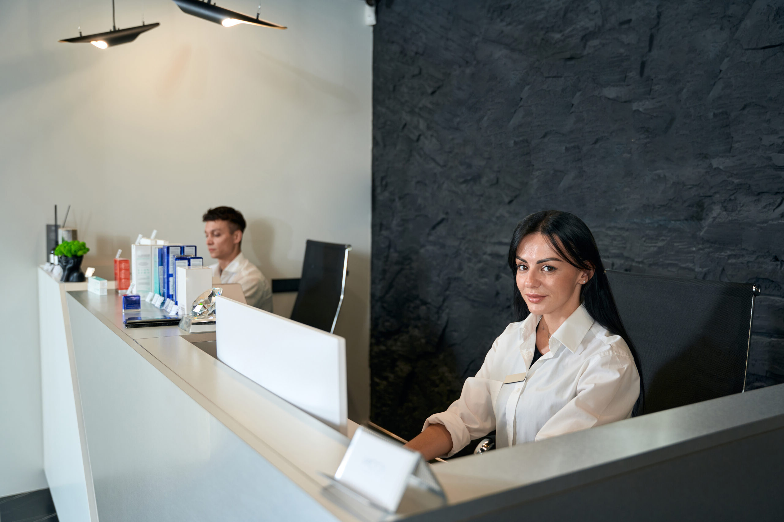 Young woman working at the computer next to her assistant, looking at the camera, waiting for clients